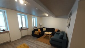 A modern living room with a blue sofa, a small table, and a yellow pouf. There are windows with white frames and a radiator on the wall. - Vacation apartment "Pfalz" - centrally located in Grünstadt (Grünstadt)