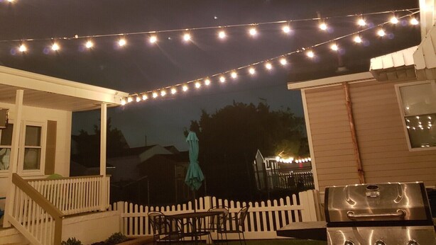 Un'area patio esterna coperta con luci a catena, un barbecue e un recinto in legno bianco.