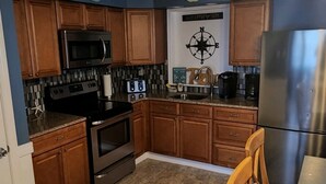 Una cucina con mobili in legno, un frigorifero in acciaio inossidabile, un forno a microonde, un forno e una macchina da caffè. Sono presenti due orologi a parete e una sedia di legno al bancone.