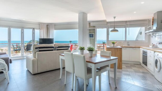 Una sala de estar moderna con sofá, mesa de comedor y sillas, una cocina con pileta y estufa, y ventanas amplias con vista al mar.