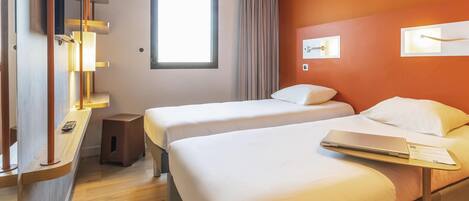 A hotel room with two beds, a desk with a laptop, a window with curtains, and a wall-mounted light fixture.