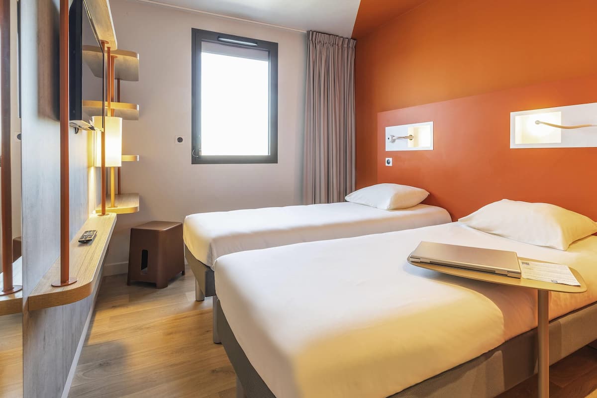 Ein Hotelzimmer mit zwei Betten, einem Schreibtisch mit Laptop, einem Fenster mit Vorhängen und einer Wandleuchte.