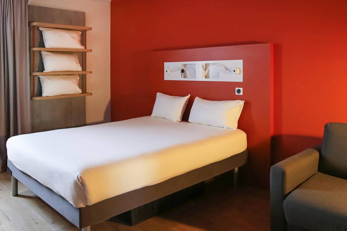 Ein Hotelzimmer mit einem Bett, einem Sessel, einem Regal mit Kissen und einem an der Wand montierten Lichtschalter.