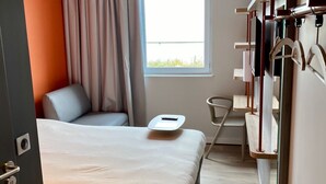 Une chambre d’hôtel moderne comprenant un lit, une chaise, une petite table, une fenêtre avec des rideaux et une armoire avec des vêtements suspendus.