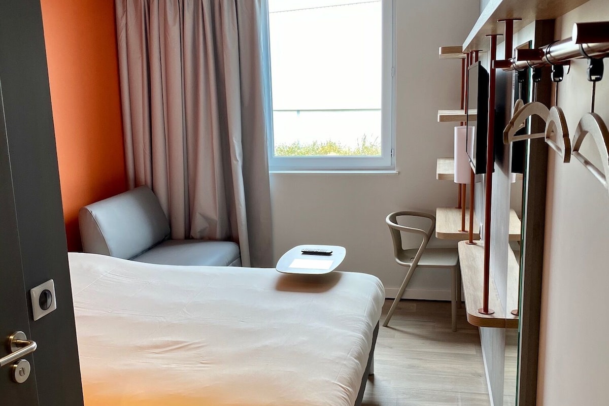 Een moderne hotelkamer met een bed, een stoel, een klein tafeltje, een raam met gordijnen en een kledingkast met daarin hangende kleding.