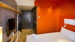 Une chambre d’hôtel équipée d’un lit, d’une télévision, d’un porte-manteau et d’une porte donnant sur une autre pièce.