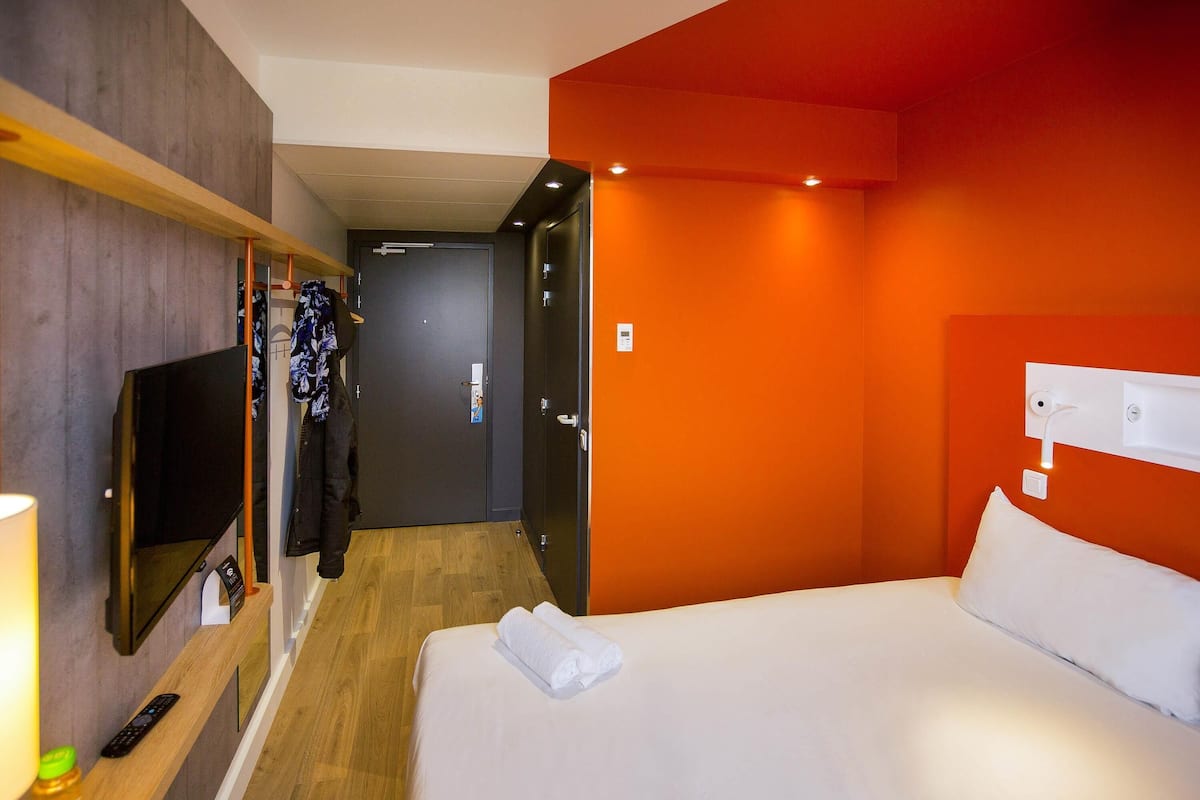 A hotel room with a bed, a TV, a coat rack, and a door leading to another room.