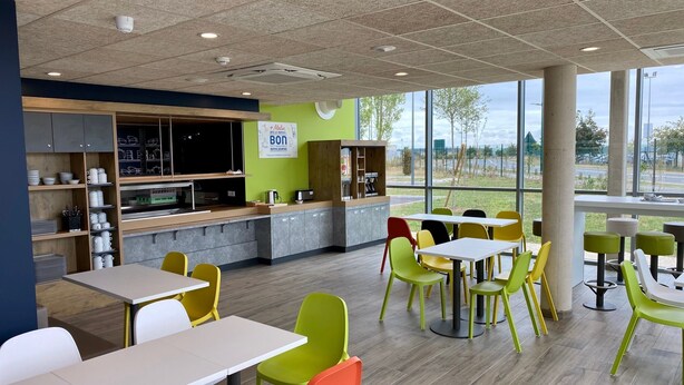 Un café moderne doté d’un espace cuisine, de chaises colorées et de grandes fenêtres donnant sur l’extérieur.