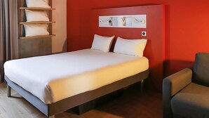 A hotel room with a bed, a chair, a shelf with pillows, and a wall-mounted light switch.