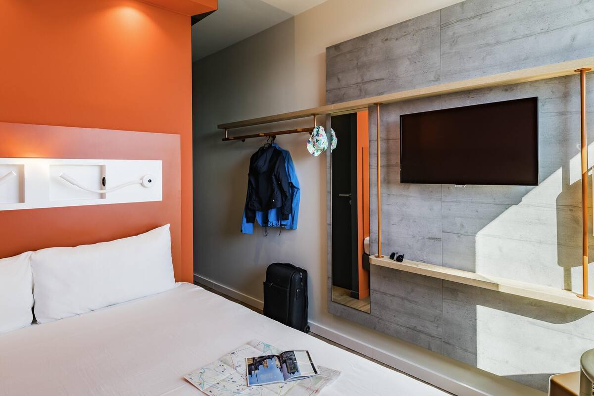 Ein Hotelzimmer mit einem Bett, einem an der Wand befestigten Flachbildfernseher, einem Kleiderständer mit Jacken und einem Koffer.