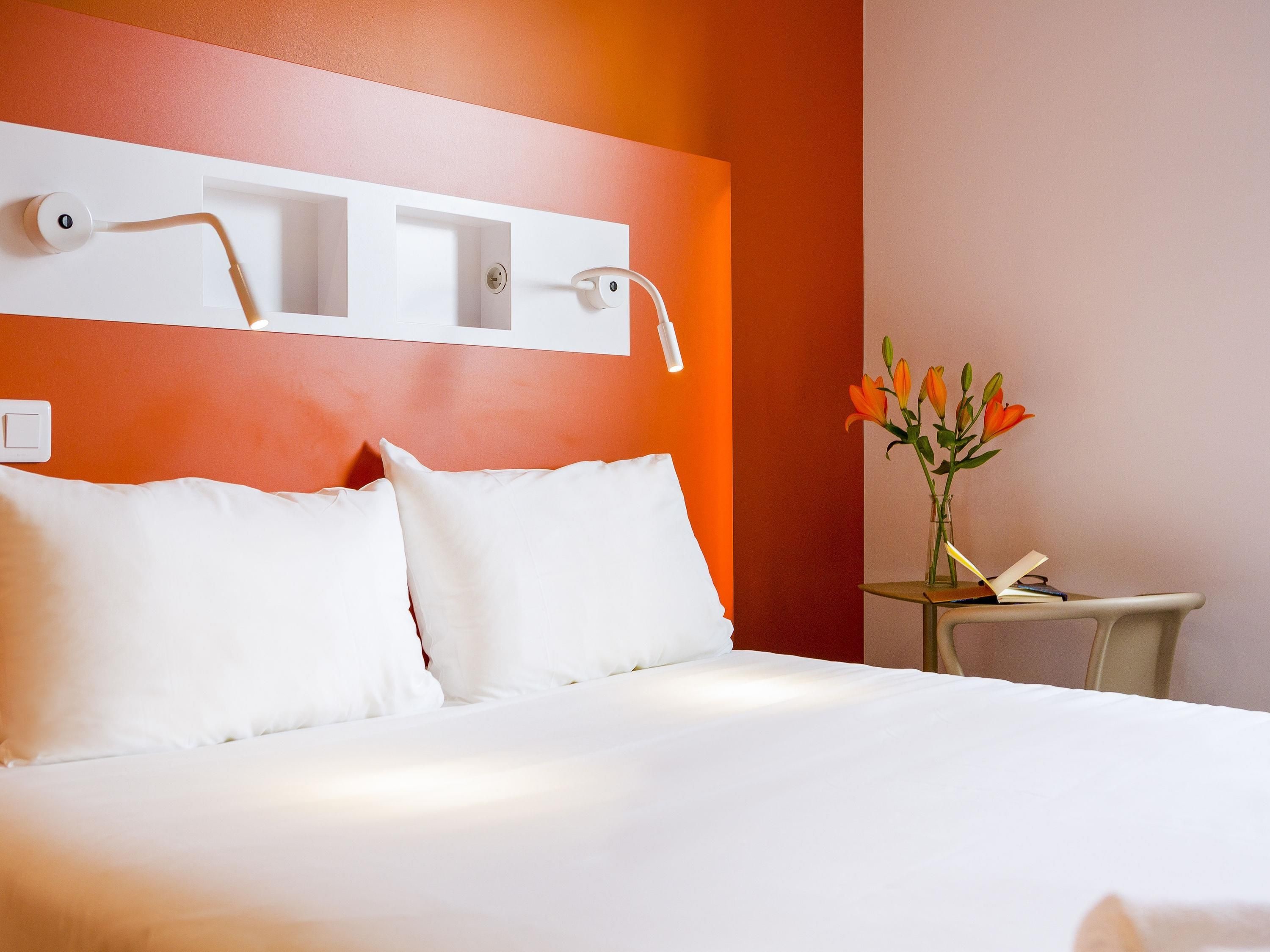 A hotel room with a bed, white pillows, a nightstand with a vase of flowers, and a wall-mounted light fixture.