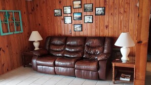 Un salón con un sofá de cuero marrón, dos mesitas de noche con lámparas, suelo de baldosas y paredes de madera con cuadros enmarcados.
