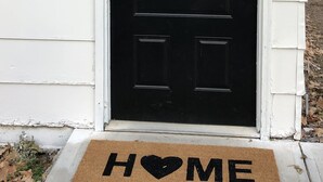 A black door with a red and black checkered bow, a black wall-mounted light, and a doormat with "HOME" written on it.