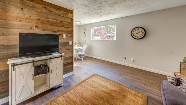 A living room with a wooden wall, a TV on a wooden cabinet, a clock on the wall, and a window.