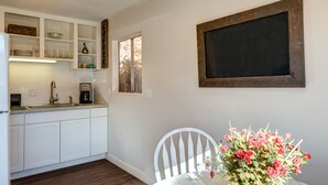 A kitchen with white cabinets, a coffee station, and a dining area with a white chair and a vase of flowers.