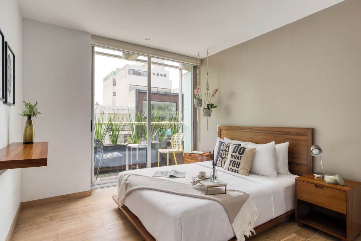 A modern bedroom with a large bed, bedside tables, a nightstand, a hanging plant, and a view of the outside.