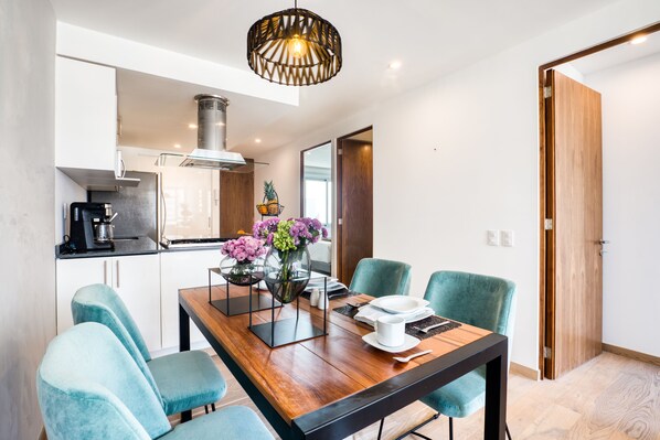 Una cocina moderna con armarios blancos, una cafetera y un comedor con mesa de madera, sillas color aguamarina y un jarrón con flores.