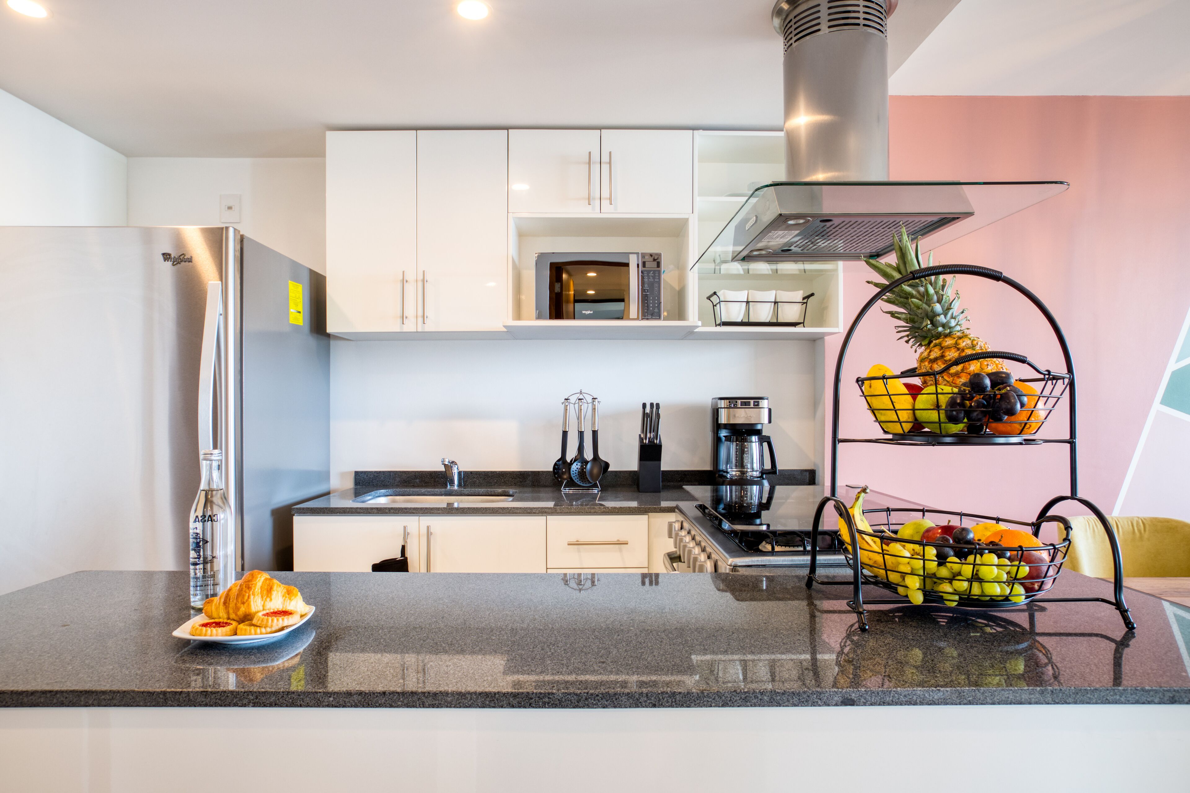 A modern kitchen with white cabinets, a stainless steel refrigerator, a coffee maker, and a fruit basket on the counter.