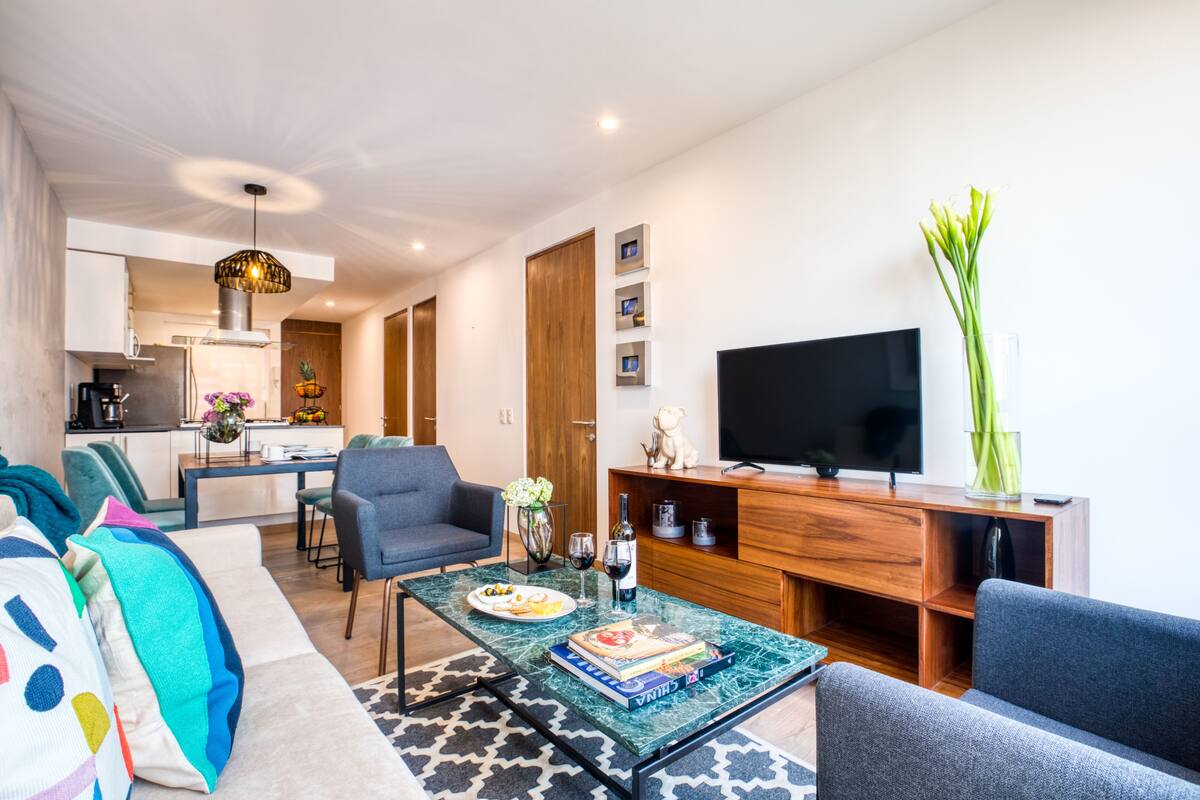 A modern living room with a sofa, coffee table, and a TV. There is a dining area with chairs and a table in the background.