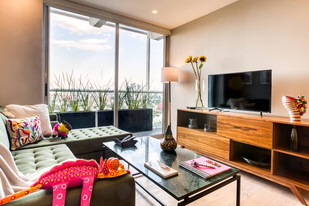 A modern living room with a sofa, coffee table, TV, and large windows overlooking a cityscape.