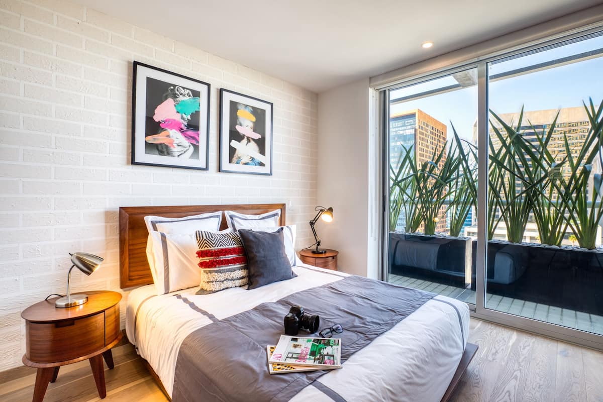A modern hotel room with a large bed, a nightstand with a lamp, a window with a view of buildings and plants, and two framed artworks on the wall.