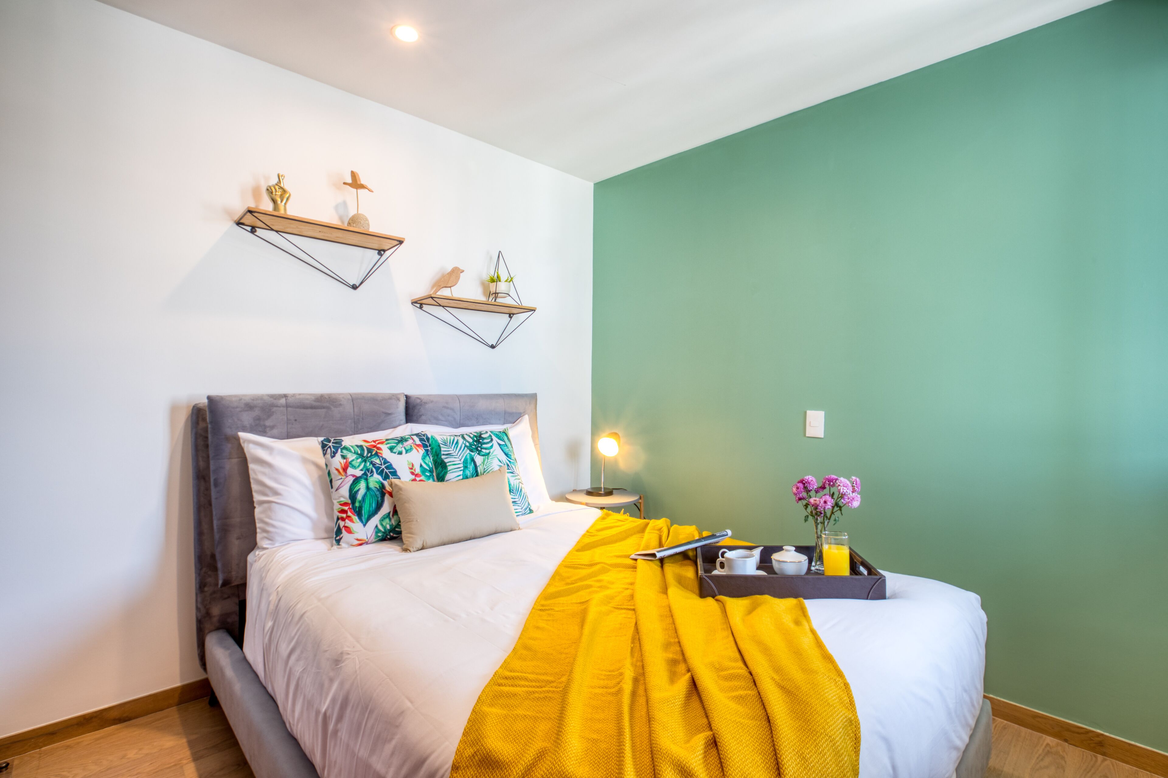 A neatly made bed with a yellow throw blanket, a bedside table with a lamp, and wall-mounted shelves with decorative items.