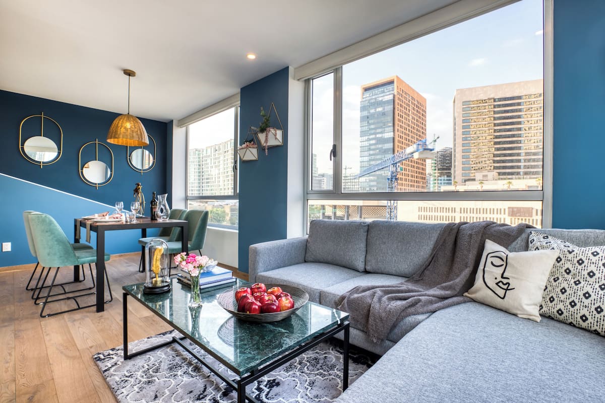 A modern living room with a sofa, dining table, and chairs, large windows with a city view, and decorative mirrors.