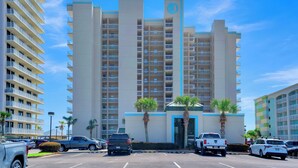 A multi-story hotel with a central entrance, parking lot, and surrounding palm trees.