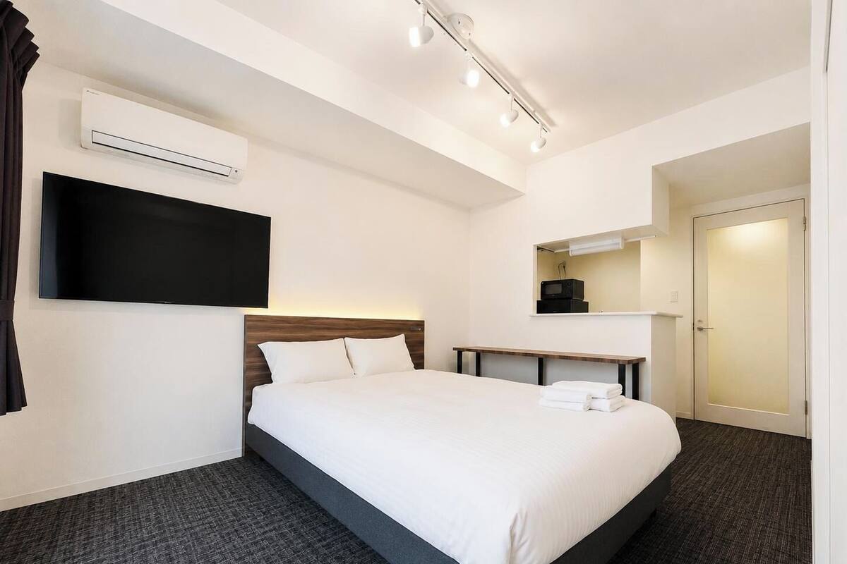 A hotel room with a bed, a flat-screen TV mounted on the wall, a bedside table, an air conditioning unit, and a door.