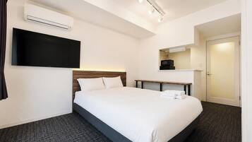 A hotel room with a bed, a flat-screen TV mounted on the wall, a bedside table, an air conditioning unit, and a door.