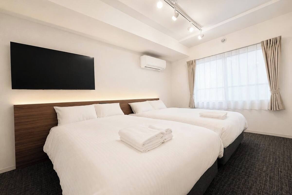 A hotel room with two beds, a flat-screen TV, an air conditioning unit, and a large window with curtains.