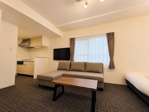 Habitación de hotel con sofá, una pequeña mesa, televisor, cama, zona de cocina y ventana con cortinas.