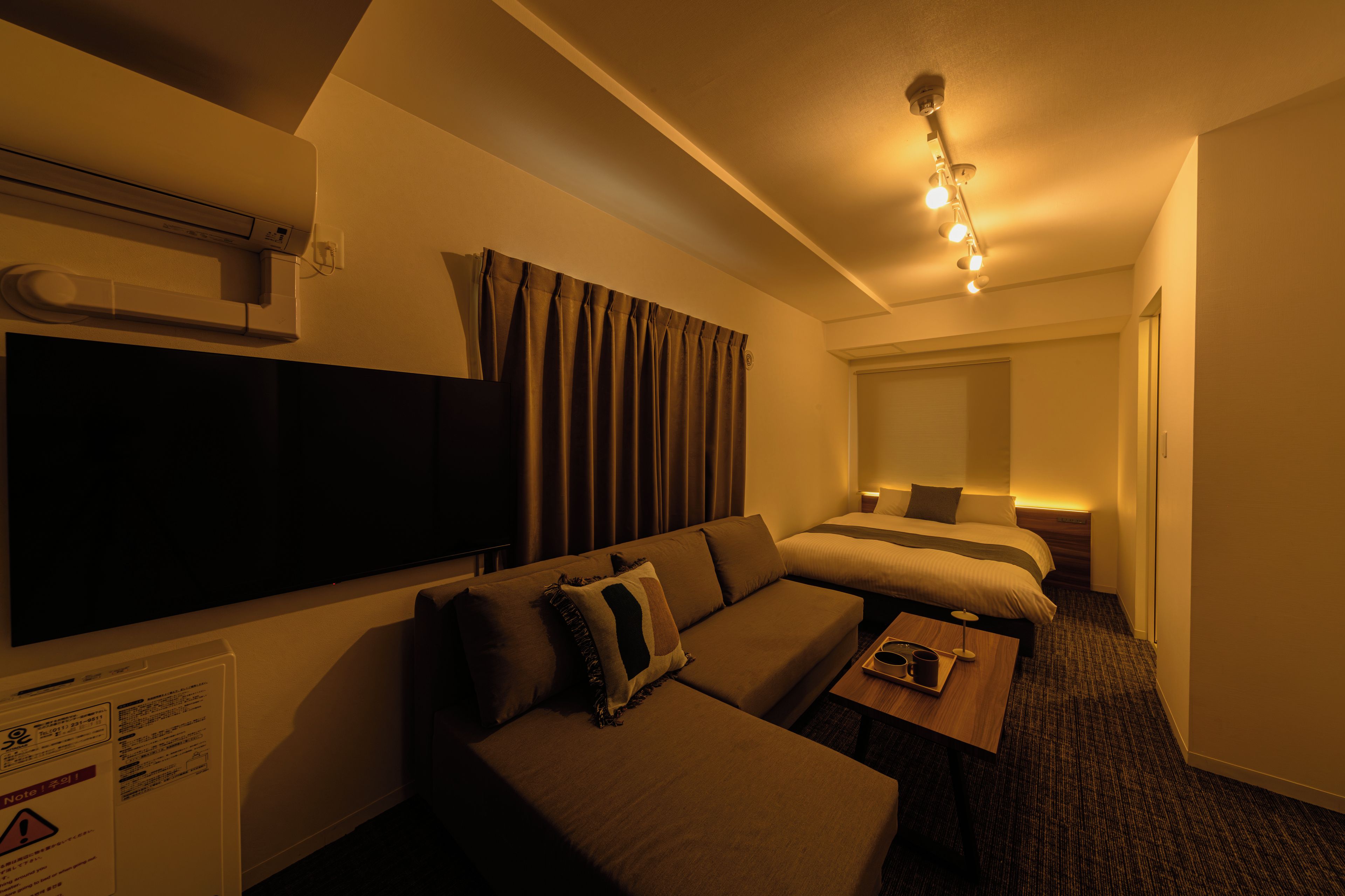 A hotel room with a sofa, a bed, a TV, and a bedside table with a lamp.