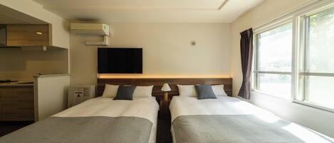 A hotel room with two beds, a TV, a window with curtains, and a kitchenette.
