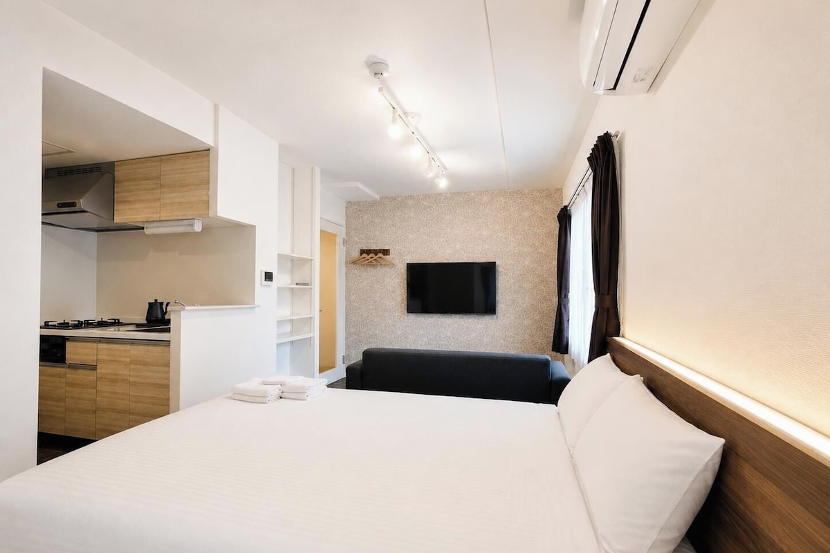 Una habitación de hotel moderna con una cama, un televisor de pantalla plana en la pared, una kitchenette con estufa y un equipo de aire acondicionado instalado en el techo.