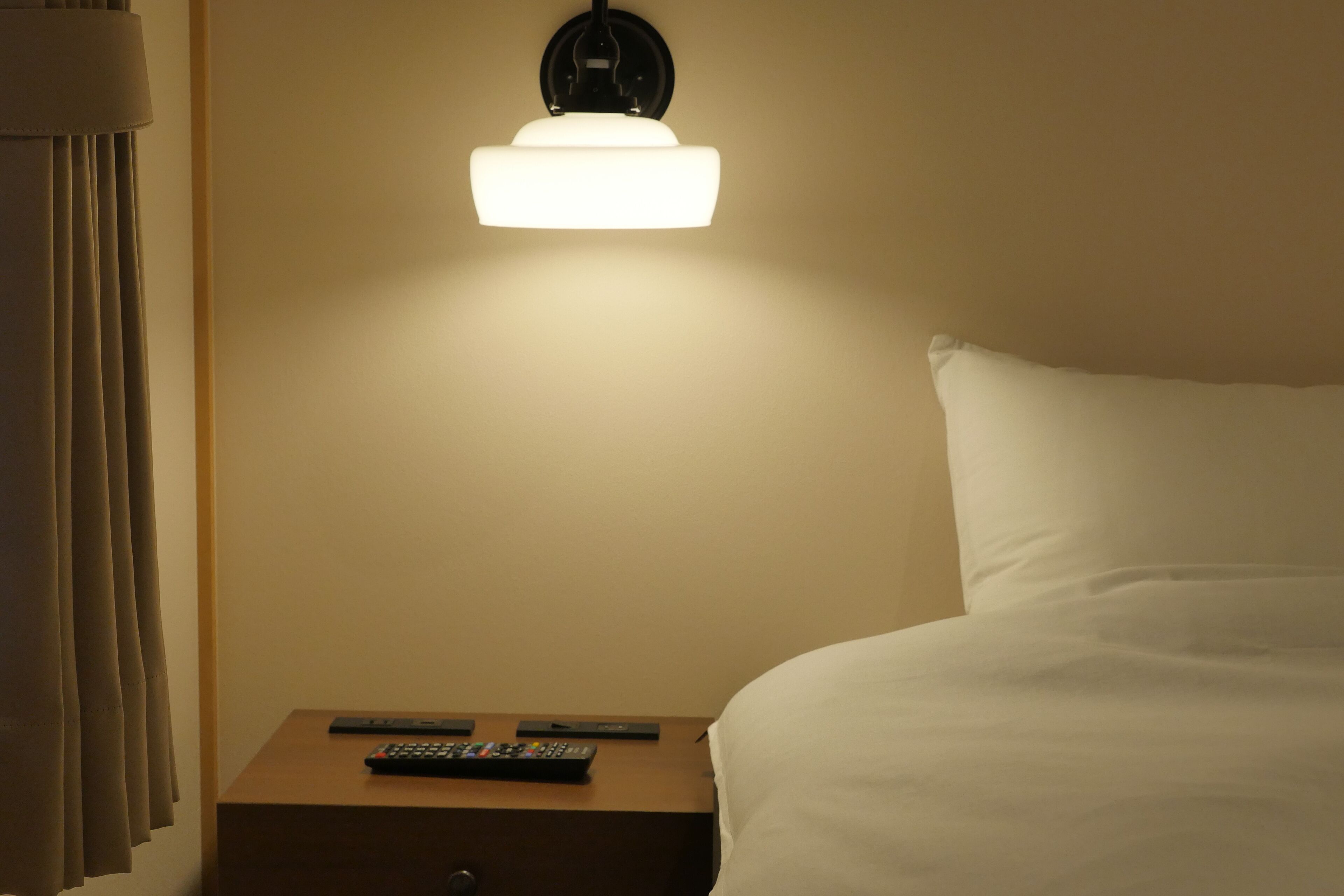 A hotel room with a bed, a nightstand with a remote control, and a wall-mounted light fixture.
