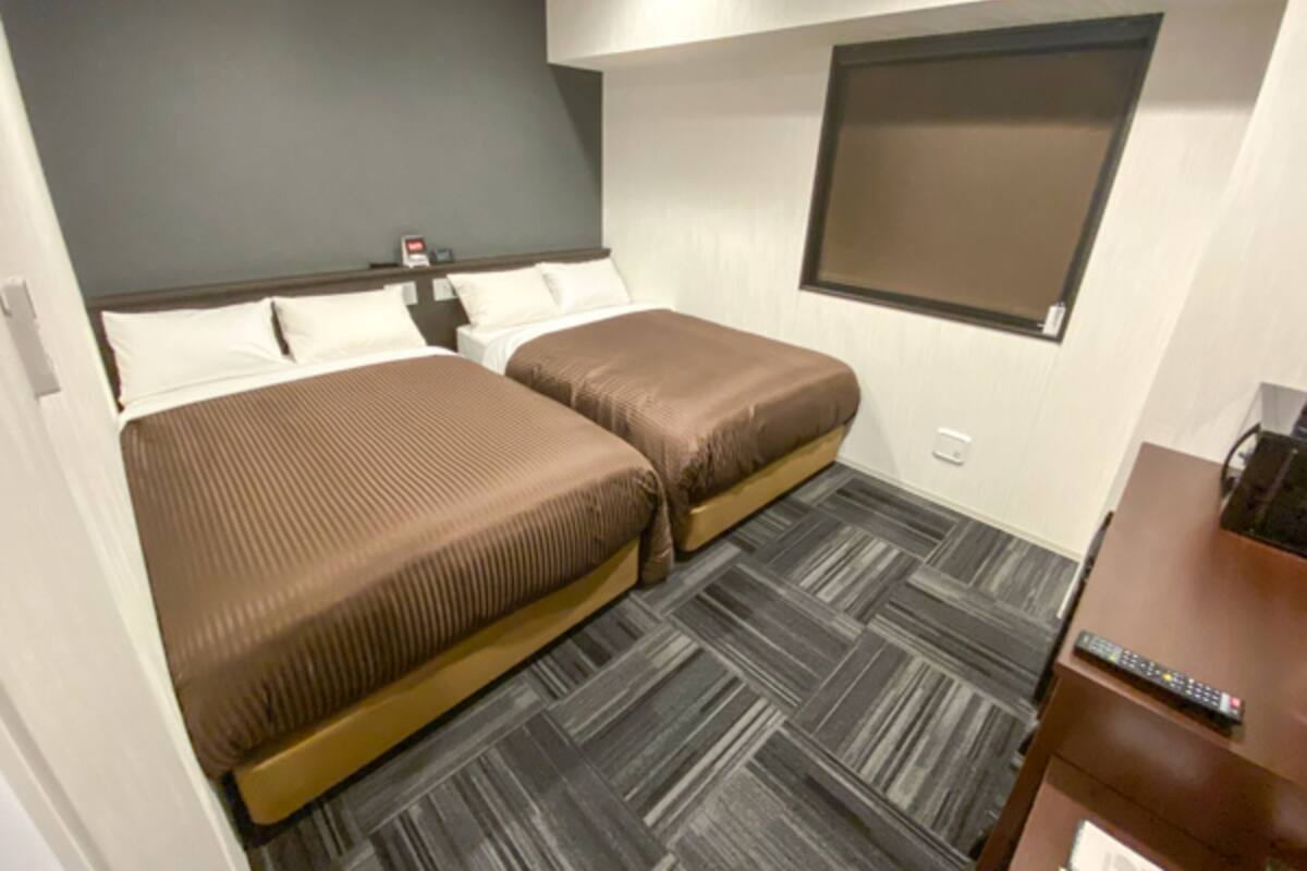 Habitació d'hotel amb dos llits, una tauleta de nit, escriptori amb un ordinador i finestra amb persianes.