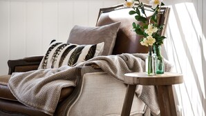 Une pièce chaleureuse avec un canapé en cuir marron, des coussins à motifs, un plaid tricoté et un vase rempli de fleurs posé sur une petite table en bois.