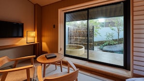 Een moderne kamer met een groot schuifglas dat uitkomt op een buitenruimte met een houten terras en een jacuzzi. Er is een televisie aan de muur bevestigd en een klein rond tafeltje met een boek.