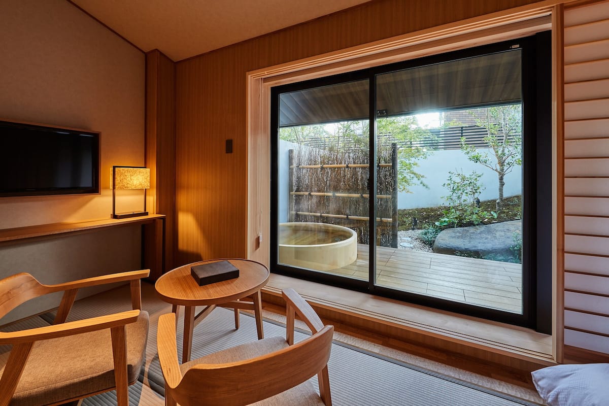 Ein moderner Raum mit einer großen Schiebeglastür, die zu einem Außenbereich mit Holzterrasse und Whirlpool führt. An der Wand ist ein Fernseher angebracht, und ein kleiner runder Tisch steht dort mit einem Buch darauf.