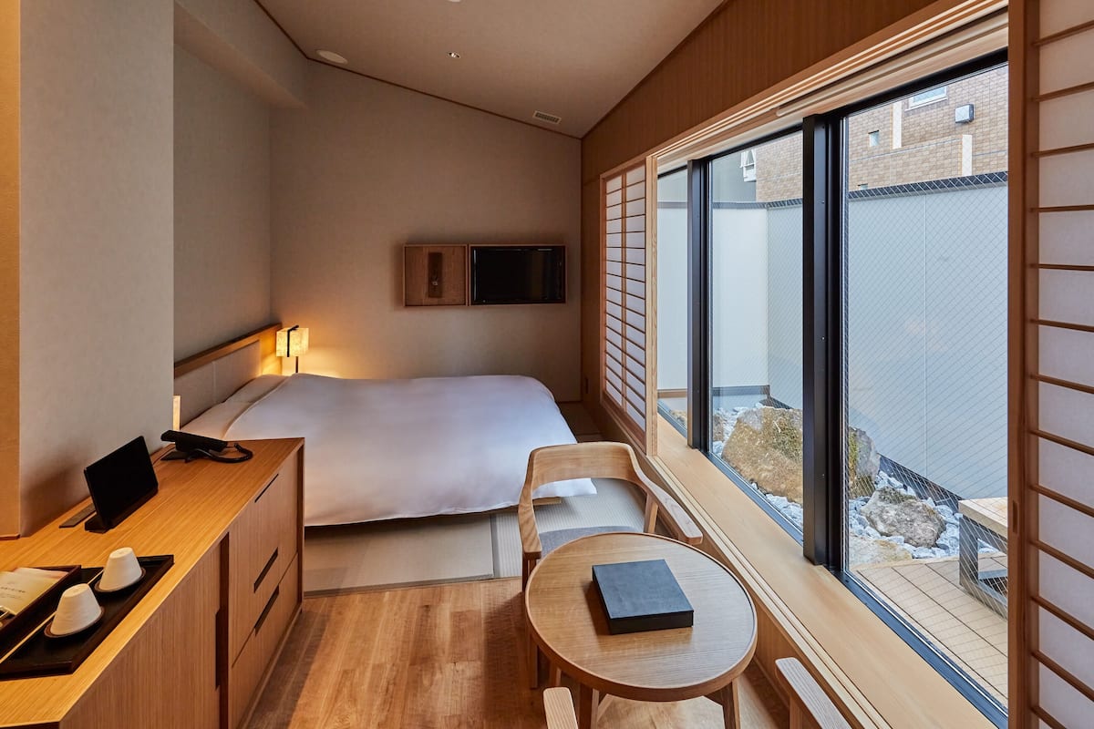 Ein modernes Hotelzimmer mit Bett, Nachttisch, Schreibtisch und Stuhl. Ein Fenster bietet Blick ins Freie.