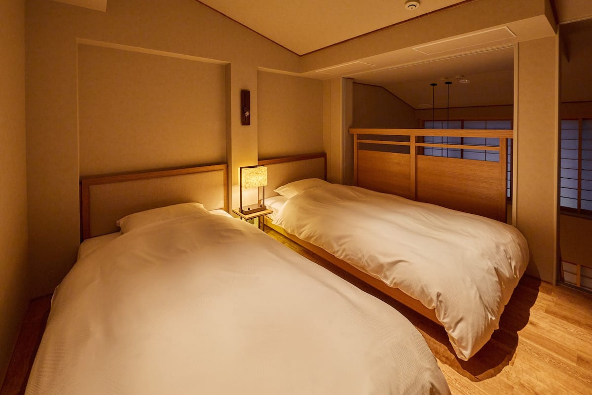 Ein Hotelzimmer mit zwei Betten, einem Nachttisch mit Lampe, Holz-Kopfteilen und Blick auf den Himmel durch ein Fenster.