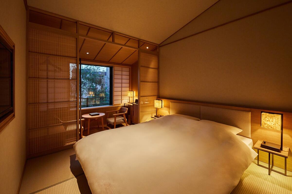 Ein Hotelzimmer mit einem großen Bett, einem Schreibtisch mit Stuhl und Blick ins Freie durch ein Fenster.