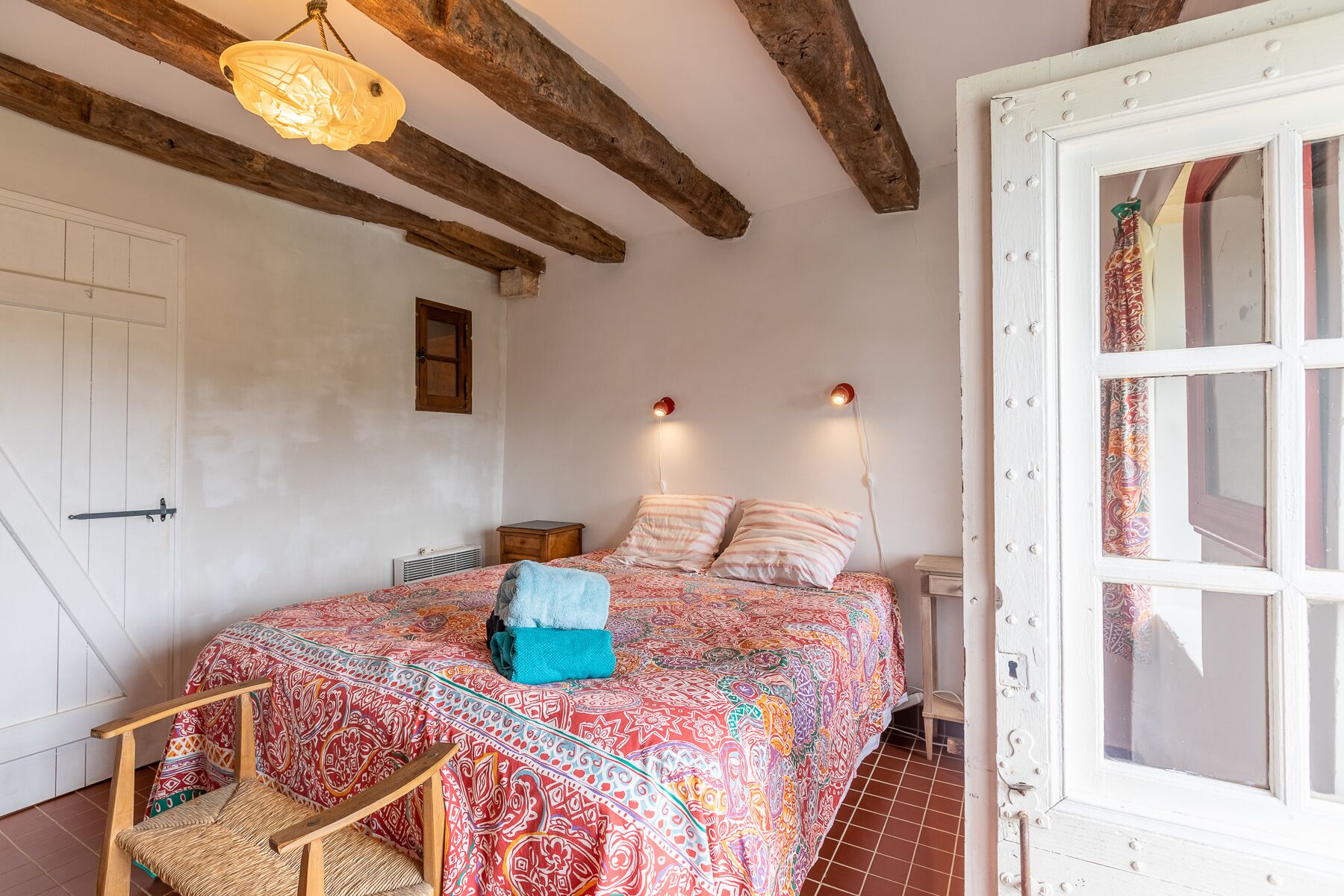 A bedroom with a bed, a wooden chair, a hanging light fixture, a window with curtains, and exposed wooden beams on the ceiling.