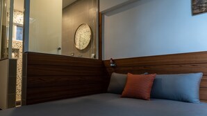 Quarto com cabeceira de madeira, cama com roupa de cama cinza e azul, e uma pequena mesa de cabeceira com um objeto vermelho.