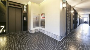 A hotel corridor with herringbone-patterned flooring, elevators, and a wall-mounted control panel. - Hotel Chicago (Changwon)
