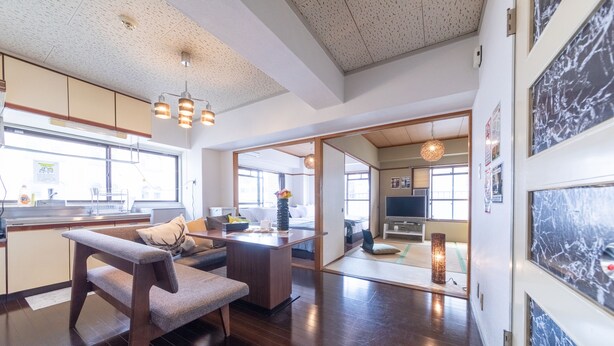 A spacious living area with a wooden bench, a sofa, and a dining table. There is a kitchen area with a sink and cabinets, and a television in the background. - SUNDAY peace room Nakashima (Hiroshima)