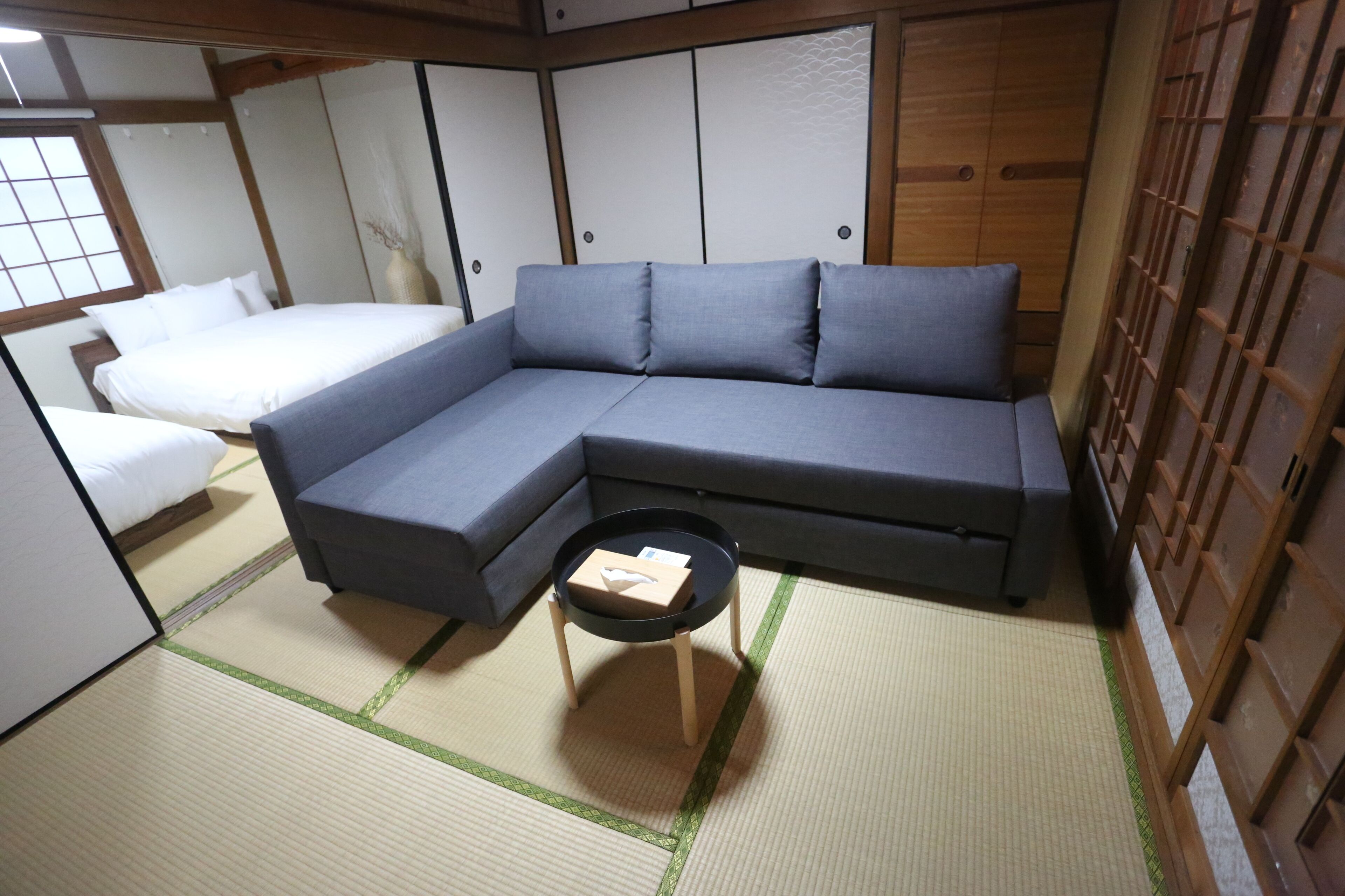A traditional Japanese-style room with tatami flooring, a grey sofa, a small wooden table, and a white bed.