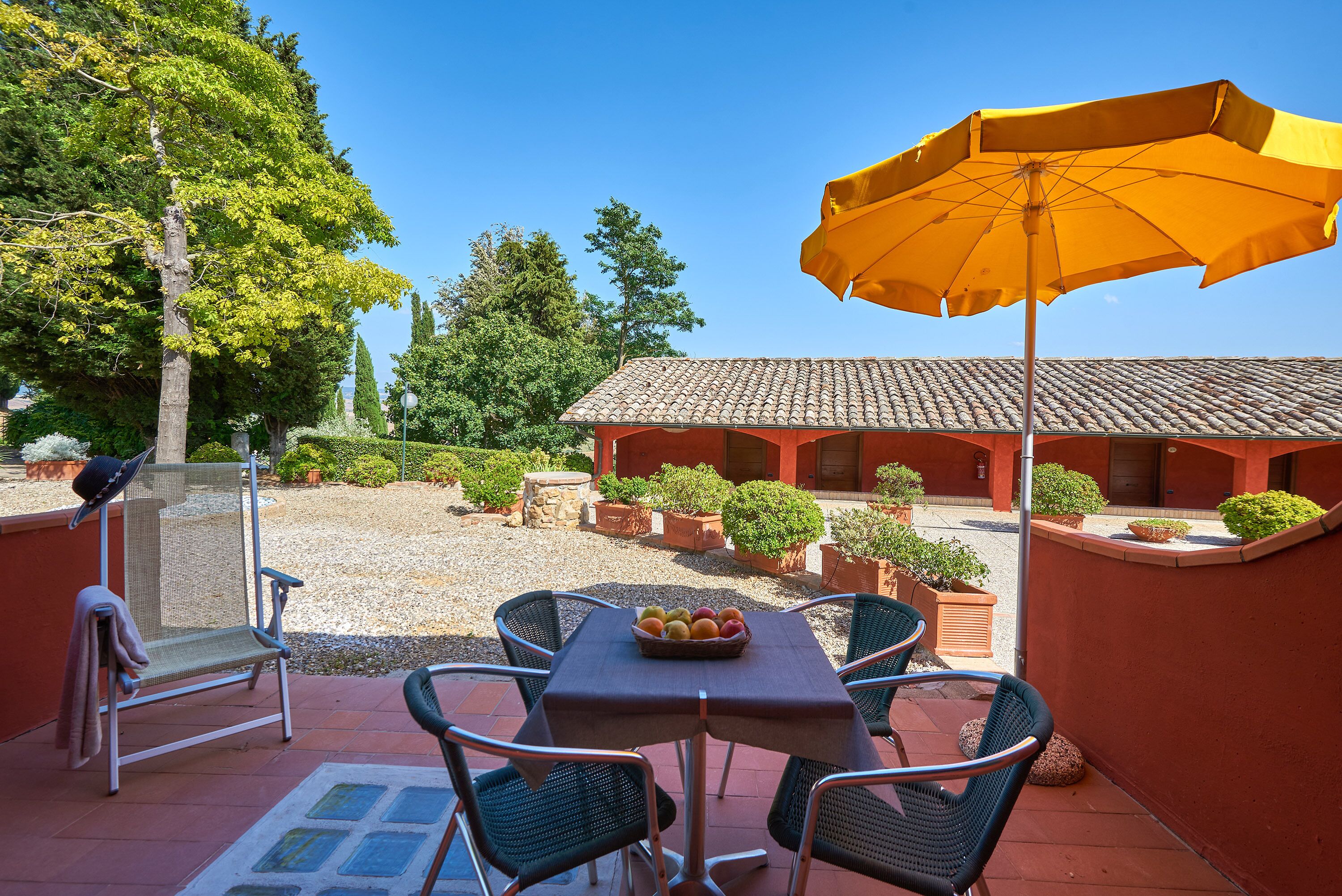 A patio with a table, chairs, and an umbrella. There is a building in the background and a gravel area with potted plants.