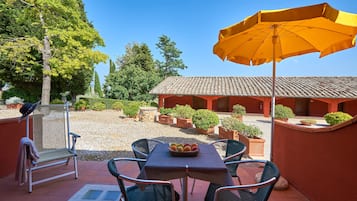 A patio with a table, chairs, and an umbrella. There is a building in the background and a gravel area with potted plants.